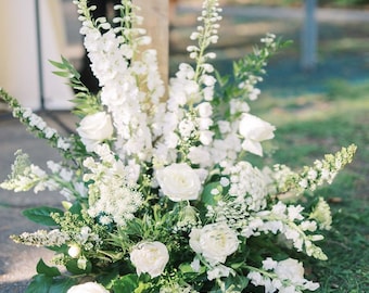 Op de afbeelding: Een weelderig bloemstuk met witte rozen, ridderspoor en andere witte bloemen. De compositie is ongeveer 51 cm lang, 41 cm breed en 51 cm hoog. Groene bladeren contrasteren met de witte bloemen. Geschikt voor bruiloften of speciale evenementen.
