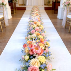 May include: A white aisle runner decorated with a colourful floral display. The flowers feature pink, yellow, and orange roses, with blue and green foliage. The arrangement is centrally placed on the runner.