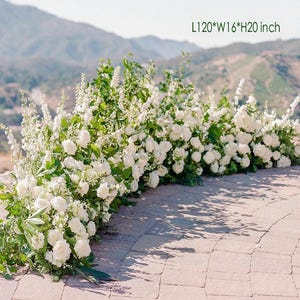 May include: A lush floral arrangement featuring white roses and greenery. The arrangement is approximately 120 inches long, 16 inches wide, and 20 inches high. The flowers are arranged on a stone surface with a mountain backdrop.