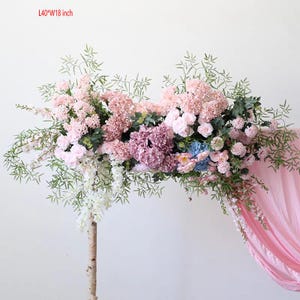 May include: A floral arch made of pink and white artificial flowers, greenery, and branches. The arch is 40 inches long and 18 inches wide.