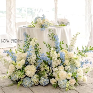 Könnte beinhalten: Ein Blumenarrangement mit weißen, hellblauen und grünen Blumen. Das Arrangement befindet sich am Fuß eines Tisches mit einer weißen Tischdecke. Die Abmessungen des Arrangements sind L101.6cm*B40.6cm*H50.8cm.