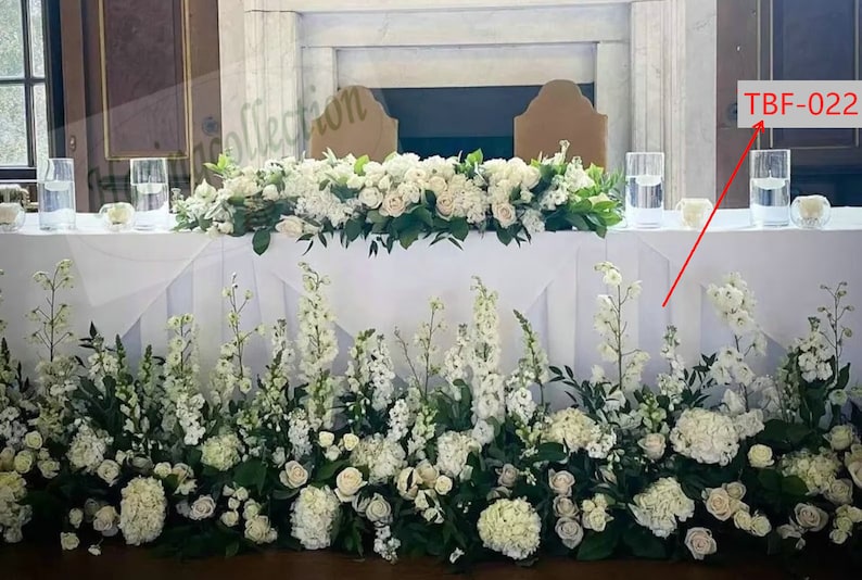 May include: A white wedding table with a long garland of white flowers and greenery. The table is set with two tall clear glass vases filled with water and white flowers. The text "TBF-022" is visible in the upper right corner of the image.