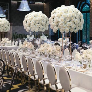 Puede incluir: Decoración elegante para una recepción de boda con largas mesas adornadas con manteles blancos, detalles dorados y grandes arreglos florales de rosas blancas. Sillas doradas y candelabros decorativos.