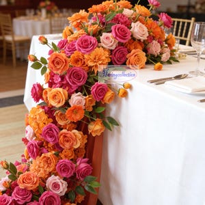 May include: A colourful floral display of roses and other flowers in shades of orange, pink, and rose cascades down a white tablecloth. Green leaves provide contrast. Cutlery and glassware are set on the table, suggesting a special occasion or event.