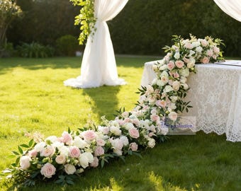 Puede incluir: Montaje para una ceremonia de boda al aire libre. Una mesa cubierta con un mantel de encaje blanco, adornada con un arreglo floral en cascada de rosas blancas y rosas, y vegetación. La escena se desarrolla en un césped verde, con un arco blanco drapeado al fondo.