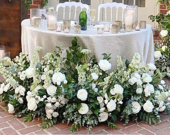 Op de afbeelding: Een ronde tafel, bedekt met een crèmekleurig tafelkleed, gedekt voor een speciale gelegenheid. Het is versierd met kaarsen, glaswerk en een groene vaas. Een weelderig bloemstuk van witte rozen en groen siert de basis van de tafel.
