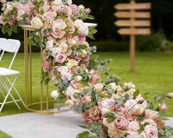 Puede incluir: Arreglo floral con rosas rosas, melocotón y crema, con follaje verde, cayendo en cascada desde un soporte de metal dorado. El arreglo está sobre una alfombra gris, con sillas blancas y un letrero de madera borroso en el fondo.