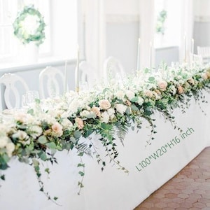 May include: A white tablecloth decorated with a floral arrangement of white and peach roses, greenery, and trailing vines. The table is 254 cm long, 51 cm wide, and 41 cm high. The setting includes white chairs and candles.