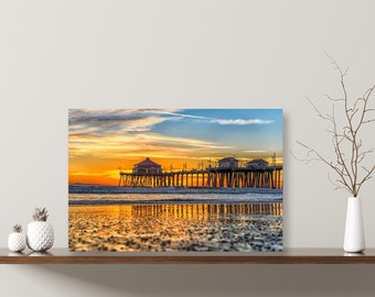 May include: A scenic photograph of a pier extending into the ocean at sunset. The sky is a vibrant orange and pink, with clouds reflecting in the water. The pier is silhouetted against the sunset, with a small building at the end.