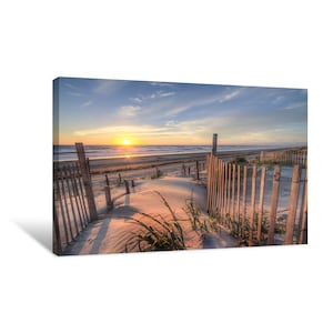 May include: A wooden fence leads to a sandy beach with a sunrise in the distance. The fence is made of vertical wooden slats and is partially obscured by tall grasses.