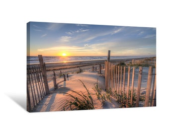 May include: A wooden fence leads to a sandy beach with a sunrise in the distance. The fence is made of vertical wooden slats and is partially obscured by tall grasses.