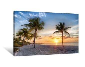 Può includere: Un tramonto pittoresco su una spiaggia con palme che si stagliano contro il cielo arancione e rosa. La spiaggia è di sabbia bianca e l'acqua è di un blu calmo.