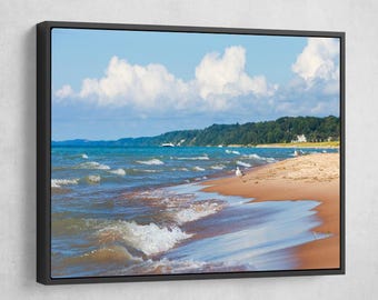 May include: A seascape photograph featuring a sandy beach with gentle waves lapping at the shore. The sky is a bright blue with fluffy white clouds. The beach is lined with green trees and a distant shoreline.