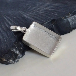 Op de afbeelding: Zilveren hanger met een rechthoekige bezel voor een steen of andere versiering. De hanger heeft een lus om aan een ketting of koord te bevestigen.