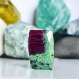 May include: A green and red gemstone with a rectangular shape. The gemstone has a green base with red streaks and black flecks.