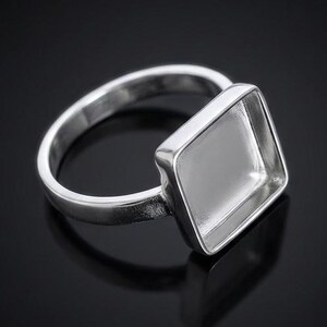 May include: A silver ring with a square bezel setting. The ring is made of a shiny, reflective metal and sits on a black surface. The square setting is empty, ready for a gemstone or other embellishment. Jewellery.