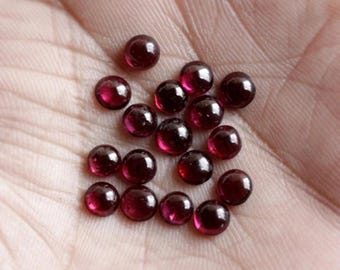 Könnte beinhalten: Eine Ansammlung kleiner, runder, polierter Edelsteine in tiefroter Farbe. Die Edelsteine sind auf einer hellen Oberfläche verteilt, wobei jeder Stein das Licht reflektiert. Die Edelsteine scheinen Cabochons zu sein, die sich für die Schmuckherstellung eignen. Jeder Edelstein hat einen Durchmesser von ca. 0,5 cm.