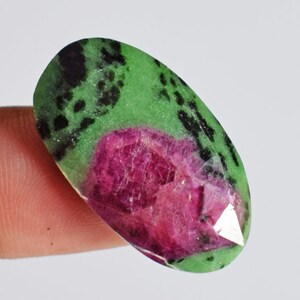 May include: An oval gemstone displaying a striking colour palette. The stone combines deep magenta and green tones, with black speckles scattered across the surface. The polished finish enhances the gem's natural beauty. A unique and eye-catching piece.