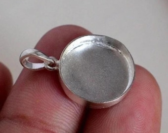 May include: A silver-colored pendant with a circular bezel and a small loop for a chain. The pendant is held in a person's hand, showcasing its polished surface and simple design. Ideal for jewelry making.