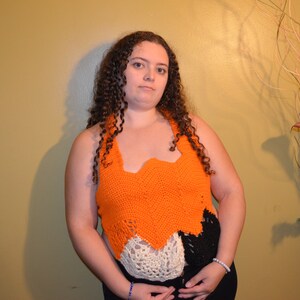 Op de afbeelding: Een vrouw draagt een gecrochete haltertop met oranje, witte en zwarte secties. De top heeft een gegolfde rand en wordt gedragen over een zwarte tanktop.
