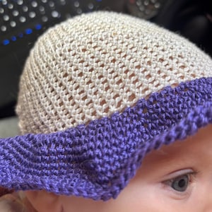 Op de afbeelding: Een crèmekleurige zonnehoed met open weefsel en een brede, paarse rand. De hoed is gemaakt van een zacht, gebreid materiaal, ideaal om de zon uit het gezicht te houden. De hoed is ontworpen voor baby's en peuters.