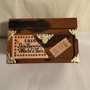 May include: A small, rectangular wooden box with a brown finish and black accents. The box features gold-colored corner brackets and a decorative gold emblem. The front of the box has text that reads "1915 San Francisco World's Fair" and a small ticket.