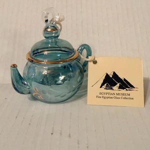 May include: A small, turquoise glass teapot with gold trim and a clear glass handle. The teapot has a matching lid and spout. A tag reads "Egyptian Museum Fine Egyptian Glass Collection."
