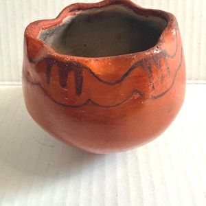May include: Hand-painted, small, orange ceramic pot with a wavy rim and dark brown decorative lines. The pot has a rounded shape and a rough, textured surface. The interior is a lighter, unglazed color.