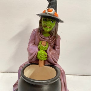 May include: A ceramic Halloween witch figurine with a green face, wearing a black hat with an orange band and a purple robe. The witch is seated, stirring a black cauldron with a wooden spoon. The cauldron is resting on a stone base.