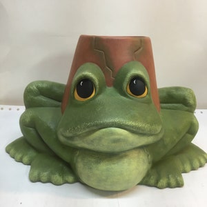May include: A green ceramic frog planter with a brown pot on its head. The frog has large, black eyes and a wide, toothy grin.