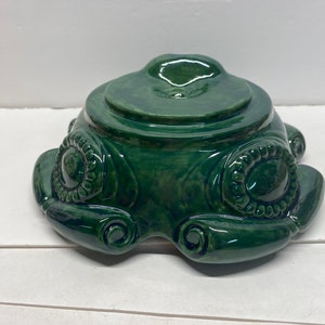 May include: A green ceramic pedestal with a round top and a decorative base. The base features a series of scrolls and circles.