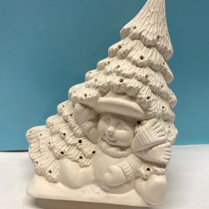 May include: A ceramic snowman figurine peeking out from behind a Christmas tree. The snowman wears a hat, scarf, and mittens. The tree has holes for lights. The figurine is unpainted and ready for customization.