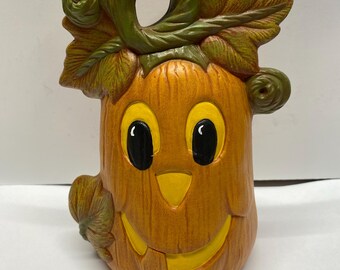 May include: A ceramic pumpkin with a friendly face, painted in orange, yellow, and black. The pumpkin has a green stem and leaves.
