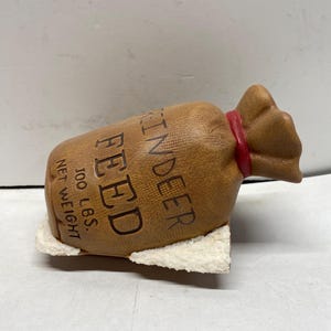 May include: A brown ceramic bag with the words "REINDEER FEED" and "100 LBS. NET WEIGHT" printed on it. The bag is tied with a red ribbon and rests on a white base. The bag is a decorative item.