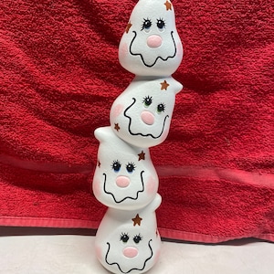 May include: A stack of three white ceramic ghost figurines with painted faces and brown stars. The ghosts are stacked on top of each other, with the largest ghost at the bottom and the smallest ghost at the top.