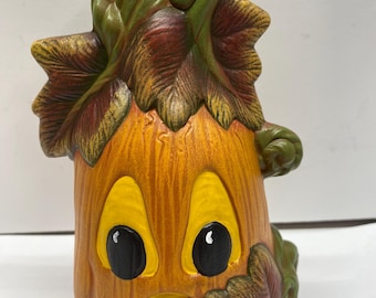 May include: A ceramic pumpkin with a green stem and leaves. The pumpkin has a painted face with yellow eyes, a black nose, and a smiling mouth. The pumpkin is a fall decoration.