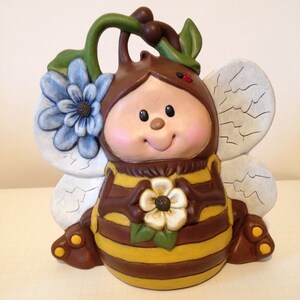 May include: A ceramic bee figurine with a brown and yellow striped body, blue flower wings, and a green stem with a red ladybug. The bee is holding a white flower in its hands.