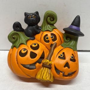 May include: A decorative ceramic figurine featuring three orange jack-o'-lanterns with carved faces, green stems, and a black cat perched on top. A small brown broom rests against the pumpkins, and one pumpkin wears a black witch hat with a purple band.
