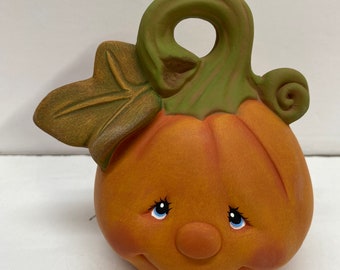 May include: A ceramic pumpkin with a green stem and leaf, painted with a smiling face and blue eyes.