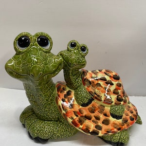 May include: A green ceramic turtle figurine with large eyes and a smaller turtle figurine riding on its back. The smaller turtle has a brown and orange shell with black spots.