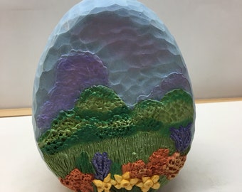 May include: A hand-carved wooden egg featuring a painted landscape scene. The egg is painted with a blue sky, purple mountains, green hills, and yellow flowers.