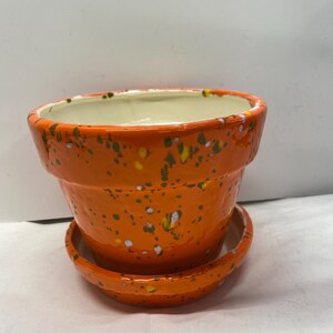 May include: Orange ceramic planter with a speckled design and a matching saucer. The planter has a white interior.