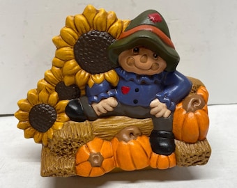 Puede incluir: Una figura de cerámica de un espantapájaros con sombrero verde y camisa azul, sentado sobre una paca de heno. La escena incluye dos grandes girasoles amarillos a la izquierda y tres calabazas de color naranja brillante en la base.