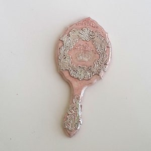 May include: A pink hand mirror with a silver ornate design featuring a crown. The mirror has a decorative handle.