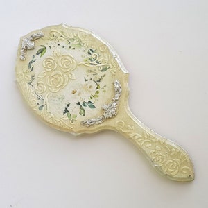 May include: A vintage hand mirror with a white and green floral design and silver accents. The mirror has a scalloped edge and a long handle.