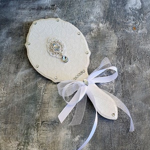May include: White hand mirror with a decorative design and rhinestones. The mirror has a white ribbon tied around the handle.