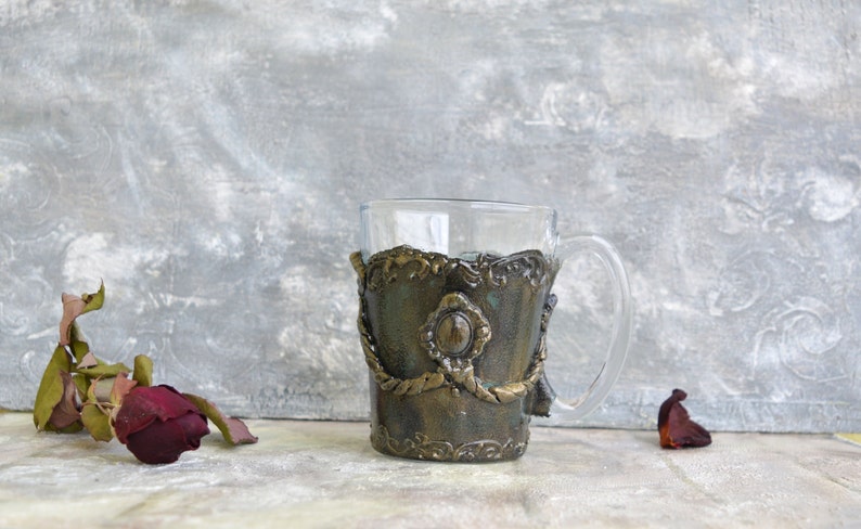 May include: A clear glass mug with a decorative gold and green clay band around the middle. The band has a raised design with a large, round, light green gem in the centre.