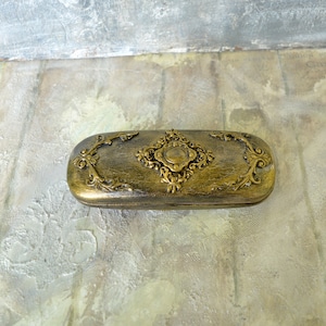 May include: A gold-colored, rectangular eyeglasses case with ornate floral designs and a central diamond-shaped embellishment.