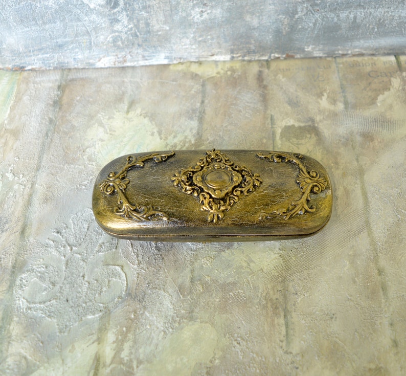 May include: A gold-colored, rectangular eyeglasses case with ornate floral designs and a central diamond-shaped embellishment.