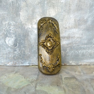 May include: A gold-colored decorative box with ornate floral designs and a raised circular center piece. The box is made of a textured material and has a slightly aged appearance.